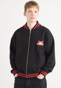 Black bomber jacket with red striped ribbed collar and cuffs, featuring an embroidered logo on the left chest, zip closure, and relaxed fit.