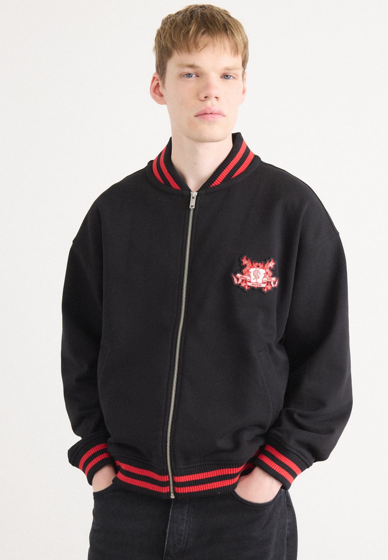 Black bomber jacket with red striped ribbed collar and cuffs, featuring an embroidered logo on the left chest, zip closure, and relaxed fit.