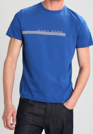 Homme portant un t-shirt bleu Hugo Boss à manches courtes et un jean foncé, une main dans la poche, sur un fond uni.