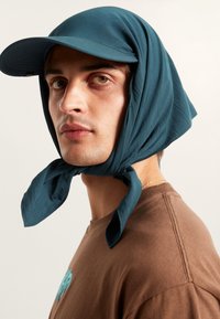 Türkiser Stoffkappe mit angeschnalltem Schal um den Hals, kombiniert mit einem braunen Shirt mit türkisfarbenem grafischen Detail. Glatte Textur, lässiges Design.