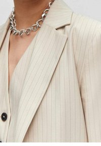Blazer beige à fines rayures avec revers crantés, fermeture par un seul bouton noir, et tissu texturé. Collier chaîne argentée visible.