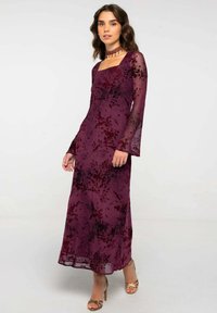 Largo vestido burdeos con escote cuadrado y mangas de campana, con un estampado floral. Hecho de tela transparente, se complementa con tacones dorados.