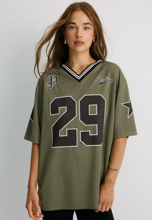 Jeune femme aux cheveux châtain clair portant un maillot de sport vert olive surdimensionné avec le numéro 29 et le texte Portland, debout devant un fond uni.