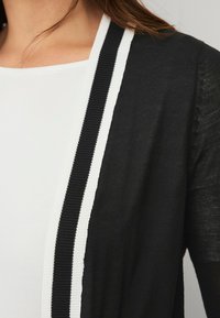 Schwarze Strickjacke mit weißen Streifenakzenten entlang der offenen Vorderseite, aus leichtem Strickmaterial mit einer leicht strukturierten Oberfläche.