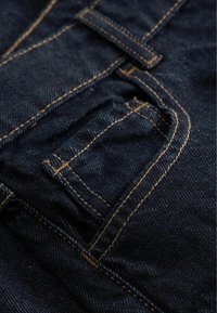 Sötétkék farmer anyag sárga varrással, kisméretű zsebbel és sima textúrával. A design hagyományos farmer stíluselemeket tartalmaz.