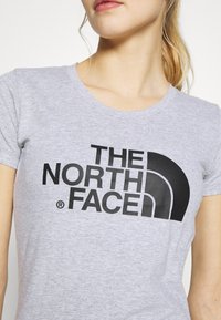 Detailní záběr na osobu oblečenou v světle šedém tričku s velkým černým logem "The North Face" na přední straně.