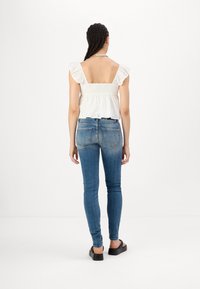Top blanco de bordado inglés con hombros con volantes, combinado con jeans ajustados de mezclilla azul y sandalias de plataforma negras. Vista trasera.