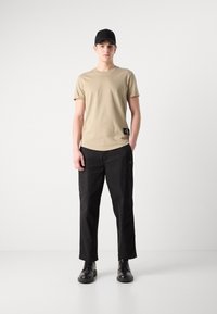 Tricou bej din bumbac, cu mâneci scurte și o etichetă cu logo, combinat cu pantaloni negri, cu croi lejer, și pantofi negri cu șiret.