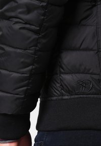 Musta untuvatakki, jossa on tikattu kuvio, joustavat hihansuut ja vyötärönauha. Takissa on hillitty kirjailtu logo alareunassa sivulla.