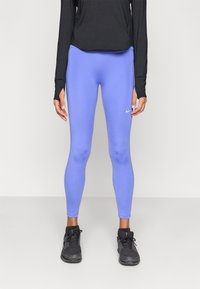 Μπλε αθλητικό κολάν από ελαστικό υλικό, εφαρμοστό σχέδιο, με μικρό λευκό λογότυπο στο αριστερό μηρό, σε συνδυασμό με μαύρα παπούτσια.