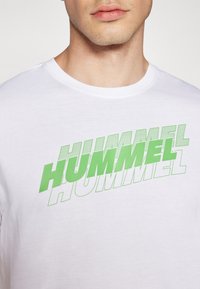 Valge puuvillane T-särk, millel on julge roheline tekst "HUMMEL" erinevates suurustes ja stiilides, sujuva tekstuuri ja ümmarguse kaelusega.
