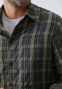 Plaid button-up shirt in green, black, and blue tones; short sleeves with a camp collar. Made from lightweight, textured fabric.