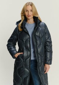 Marineblauer Steppmantel mit Kapuze, ausgestattet mit einem gesteppte Design, Reißverschluss und seitlichen Taschen. Getragen über einem leichten Pullover und Jeans.