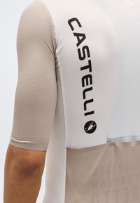 Κοντινό πλάνο μανικιού ποδηλατικής φανέλας σε μπεζ και λευκό χρώμα με υφή, με το λογότυπο "CASTELLI" και το έμβλημα σκορπιού στην πλευρική πλευρά.