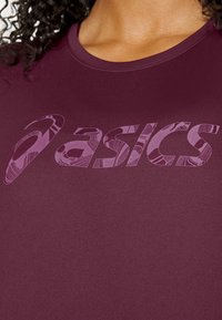 Top deportivo color marrón oscuro con mangas cortas, que presenta un gran logo "ASICS" de patrón purpúreo. Textura suave, diseño de cuello redondo.