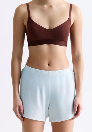 Frau trägt ein braunes Bralette mit dünnen Trägern und hellblaue elastische Shorts mit elastischem Bund, steht vor einem schlichten hellen Hintergrund.