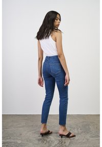 Jeans de mezclilla azul de cintura alta, con corte ajustado, dobladillo deshilachado y dos bolsillos traseros. Combinados con una camiseta sin mangas blanca y sandalias deslizantes marrones.