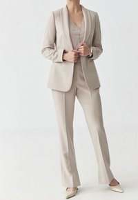 Costume beige clair sur mesure avec un blazer structuré, un pantalon assorti et un haut doux. Comprend des revers nets et des chaussures élégantes et pointues.