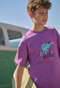 Paarse katoenen t-shirt met een blauwe chameleon graphic op een scooter. De tekst luidt "KEEP-ROLLIN'". Een jongen staat met een groen-gestreepte skateboard.