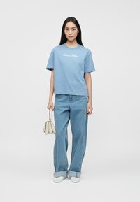 Sieviete stāv, tērpusies zila Tommy Hilfiger T-kreklā, brīvās zilos džinsos ar apkakli, baltās sporta kedās un turi mazu bežīgu rokassomu.