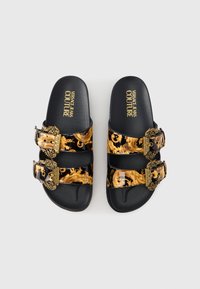 Crne natikače s debelim zlatnim baroknim uzorcima, dvostrukim remenčićima s ukrasnim zlatnim kopčama i teksturiranom tabanicom za udobnost.