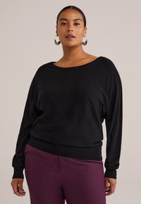 Un pull noir oversize avec des manches longues et un col large, fabriqué dans un tissu doux, associé à un pantalon violet.