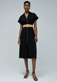 Schwarzes Hemdkleid mit kurzen Ärmeln, ausgestattet mit einem gewebten Gürtel in Taillenlänge und einem Schlitz vorne. Glattes Material und lässige Silhouette.