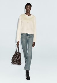 Donna che cammina indossando un cardigan avorio con bottoni, jeans blu, stivali neri e portando una grande borsa a tracolla in pelle marrone scuro.