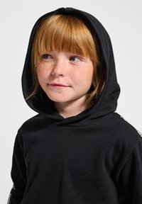 Schwarzer Kapuzenpullover aus weicher Baumwolle, mit lockerer Passform und leichten texturalen Akzenten an den Ärmeln. Sichtbare Sommersprossen auf der Haut.