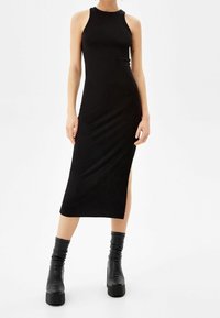 Robe débardeur noire côtelée avec un col montant et une fente latérale, associée à des bottines à plateforme noires. Texture lisse, silhouette ajustée.
