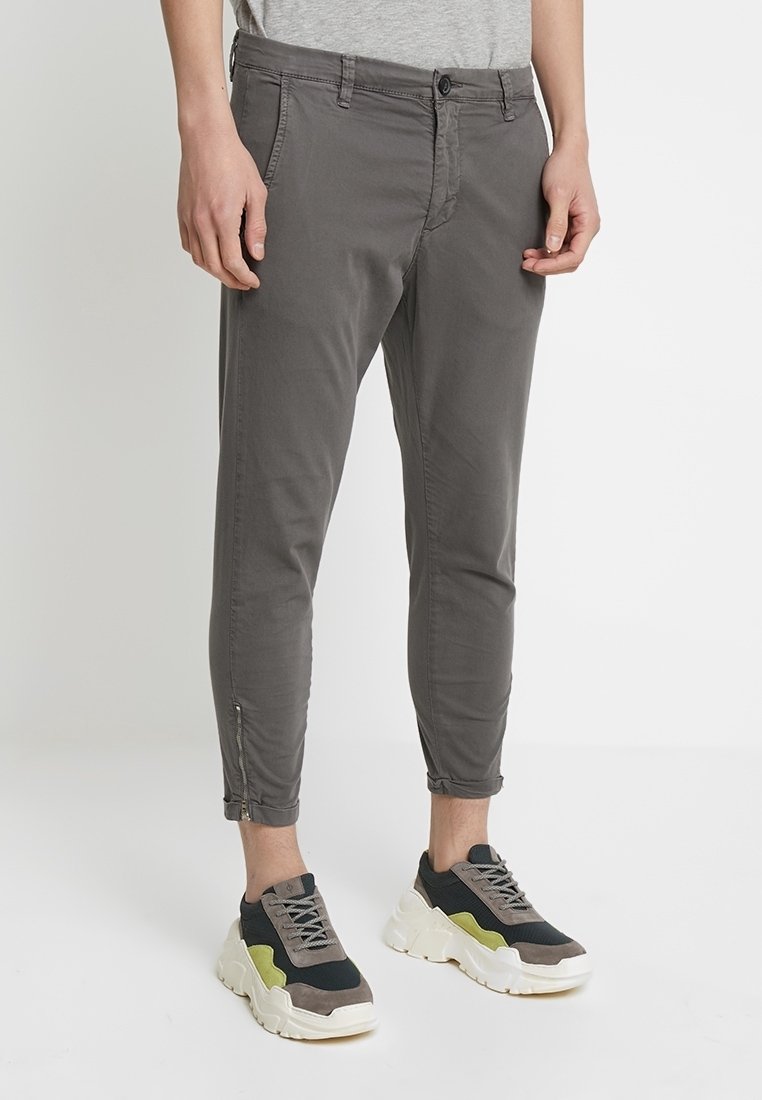 Personne portant un pantalon gris coupe slim avec des fermetures éclair à la cheville et des baskets épaisses noires, grises et vertes, debout devant un fond blanc.