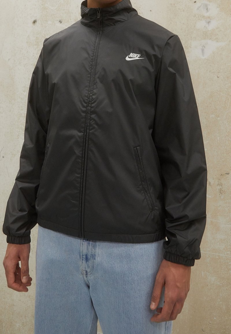 Svart vindjacka med hög krage, framdragkedja, elastiska ärmslut och sidofickor. Har en vit Nike-logotyp på bröstet.