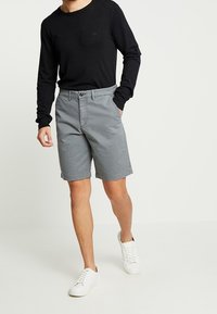 Mann in schwarzem Langarmhemd, grauen knielangen Shorts und weißen Sneakers, der mit einer Hand in der Tasche vor einem weißen Hintergrund steht.