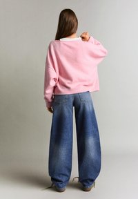 Femeie cu păr lung și castaniu, purtând un pulover roz larg și blugi evazați de culoare albastru deschis, stând desculță pe fundal simplu.