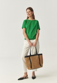 Top verde cu mânecă scurtă, pantaloni crem și balerini negri; poartă o geantă de umăr mare, împletită, de culoare piersică cu accente negre și textură detaliată.