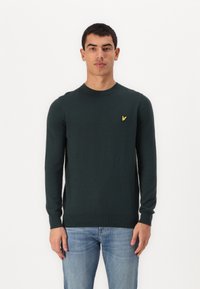 Dark green sweater with a crew neck and long sleeves. Features a small yellow embroidered logo on the chest. Smooth texture and fitted design.