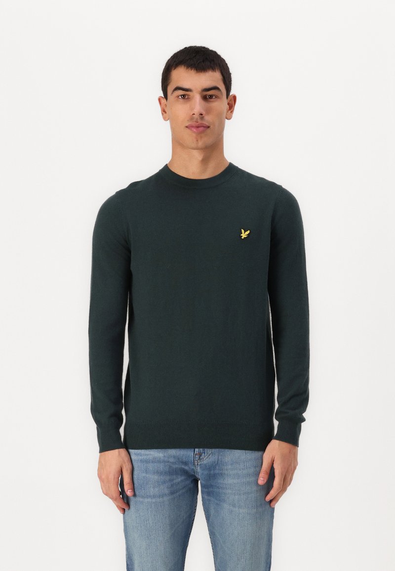 Dark green sweater with a crew neck and long sleeves. Features a small yellow embroidered logo on the chest. Smooth texture and fitted design.