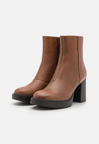 Botas de tobillo de cuero marrón con un acabado suave, tacón cuadrado alto, cremallera lateral y diseño de punta redonda. Suela de goma negra texturizada.