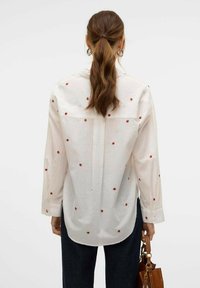 Camicia bianca con ricami floreali rossi, maniche lunghe e vestibilità rilassata. Presenta uno spacco posteriore e un orlo leggermente curvo.