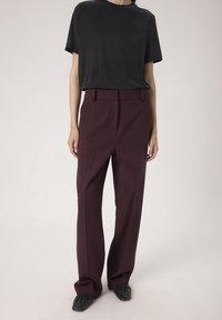 Schwarzes Kurzarm-T-Shirt kombiniert mit hoch taillierten, weit geschnittenen burgunderfarbenen Hosen, die eine glatte Textur und einen maßgeschneiderten Schnitt aufweisen. Schwarze Schuhe runden das Outfit ab.