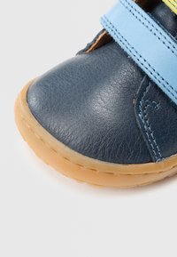 Pantof din piele albastru închis cu vârf rotund, prevăzut cu o curea Velcro albastru deschis și talpă din cauciuc. Cusături vizibile pe partea superioară.