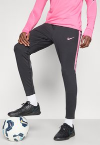 Μαύρο αθλητικό παντελόνι με ροζ λωρίδες στο πλάι, κατασκευασμένο από ελαστικό ύφασμα. Συνδυάζεται με μαύρα ποδοσφαιρικά παπούτσια και μια μοτίβο λευκή μπάλα.