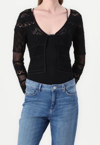 Czarny dziergany kardigan z przezroczystymi, wzorzystymi rękawami, dekoltem w serek i dopasowanym krojem. Do tego niebieskie dżinsy z jasnym wypraniem i kieszeniami.