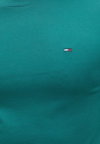 Τυρκουάζ ύφασμα με μικρό ορθογώνιο λογότυπο που περιέχει τρεις οριζόντιες ρίγες σε λευκό, ναυτικό μπλε και κόκκινο στην περιοχή πάνω στο στήθος.