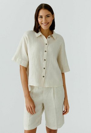 Vrouw glimlachend, draagt een beige korte mouwen overhemd met knopen en bijpassende knielange shorts, staand tegen een effen lichte achtergrond.