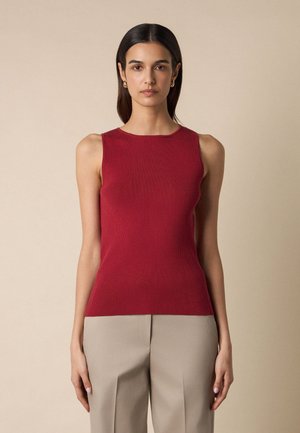 Kobieta o ciemnych włosach, nosząca złote kolczyki-koła, czerwoną, bezrękawnikową prążkowaną bluzkę i beżowe spodnie z wysokim stanem na jednolitym, beżowym tle.