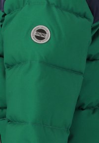 Grön puffjacka med en slät, matt yta och en rund silverlogotyp med texten "ZIGZAG", placerad på bröstet.