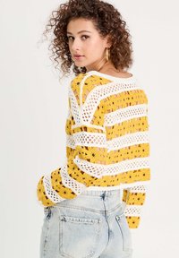 Sweater au crochet présentant des rayures jaunes et blanches, des motifs floraux et un design ajouré ; assorti à un jean en denim taille haute.