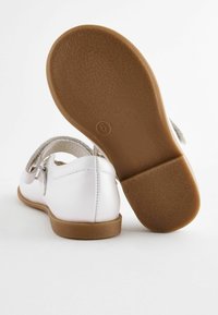 Weiße Lederschuhe mit glatter Oberfläche, ausgestattet mit einem schmalen Riemen und einer Schnallendetails. Braune Gummisohle mit strukturiertem Grip-Muster.