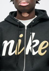 Čierna mikina s kapucňou na zips s veľkým bielym a zlatým logom „nike“. Má kapucňu s nastaviteľnými šnúrkami a viditeľný detail zipsu. Jemná textúra.